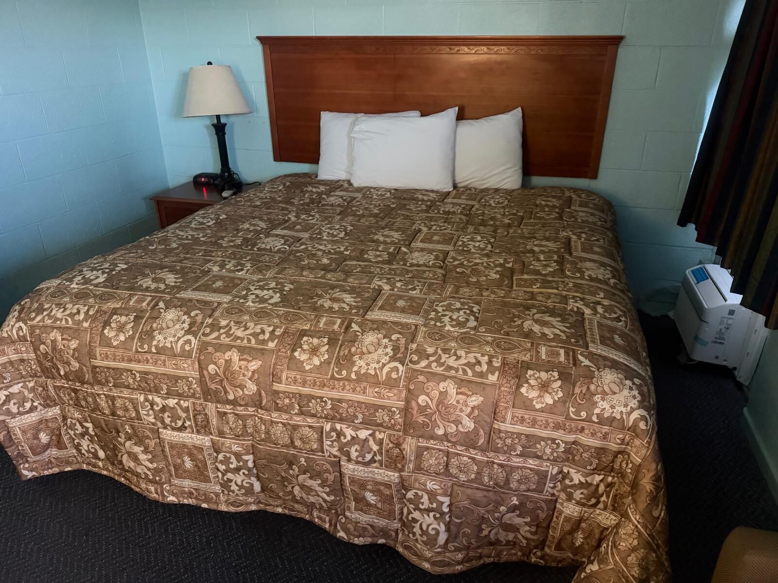 A hotel room with a large bed, a wooden headboard, a nightstand with a lamp, and a heater.