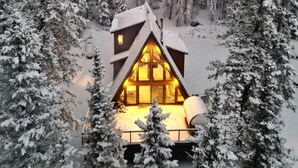 Une cabane entourée d’arbres couverts de neige, dont l’intérieur est éclairé d’une lumière chaleureuse visible à travers les fenêtres.
