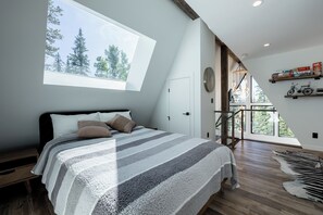 Une chambre avec une grande fenêtre, un lit avec une literie à rayures, un plancher en bois et une vue sur les arbres à l’extérieur.
