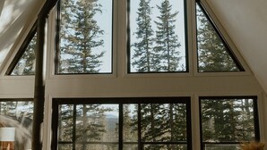 Une pièce spacieuse avec de grandes fenêtres donnant sur de grands arbres, un plafond haut avec une toiture inclinée et une cheminée.