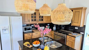 Cocina con armarios de madera, un lavamanos de acero inoxidable, una estufa y un microondas. Se observan lámparas colgantes de mimbre, un frutero y un jarrón con flores sobre la encimera.