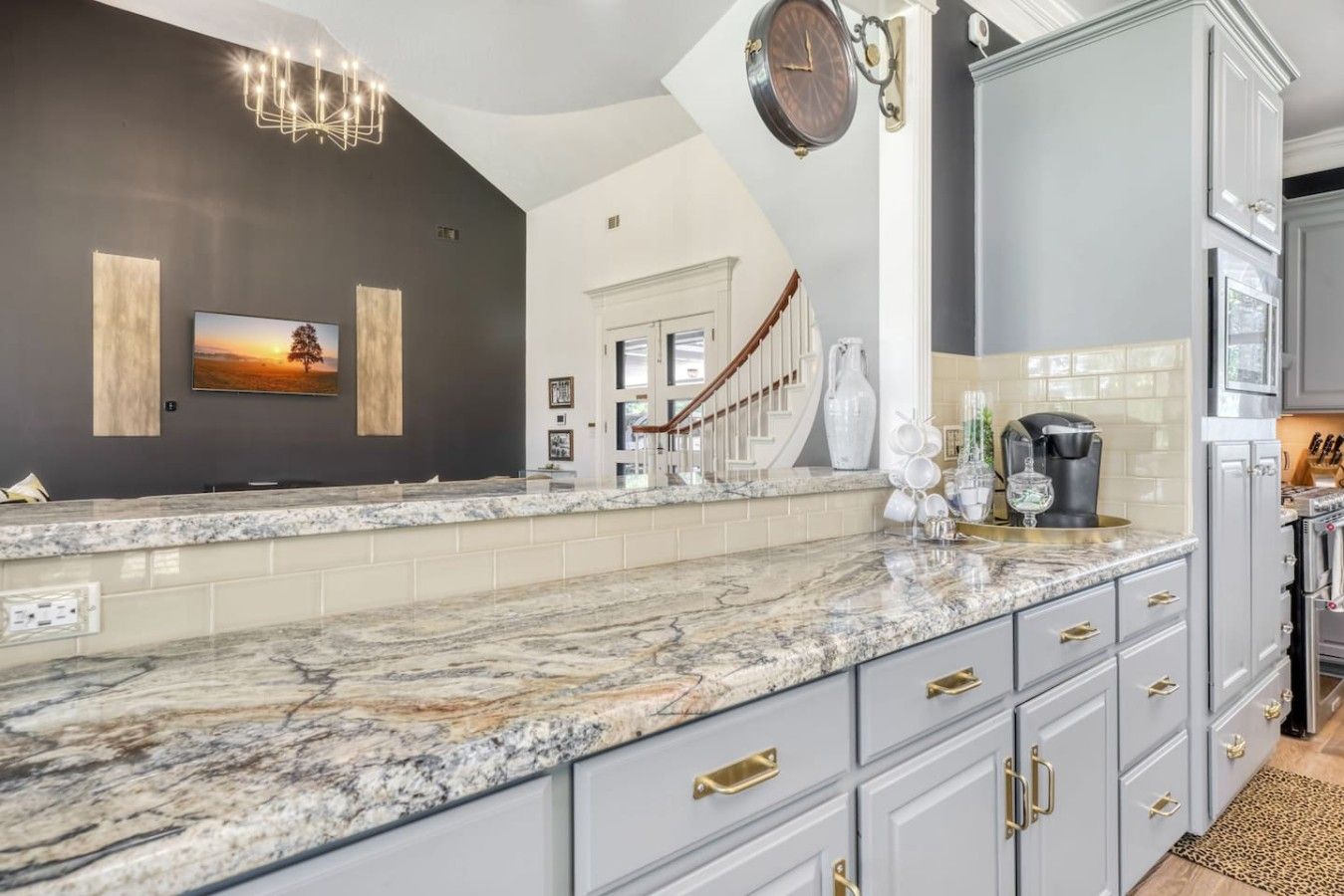 A kitchen with a marble countertop, grey cabinets, a clock, and a staircase leading to another room.