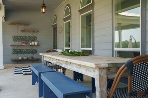 Un patio cubierto con una mesa y bancos de madera, rodeado de ventanas amplias y estantes con plantas.