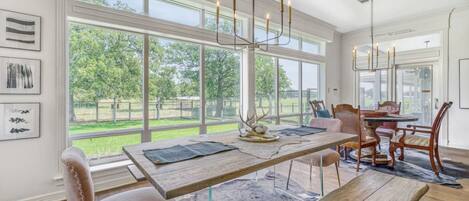 Un comedor con una mesa de madera, sillas y una araña. Amplias ventanas ofrecen vistas al exterior.