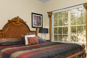 A bedroom with a wooden headboard, a bed with striped bedding, a bedside lamp, and a window with blinds.