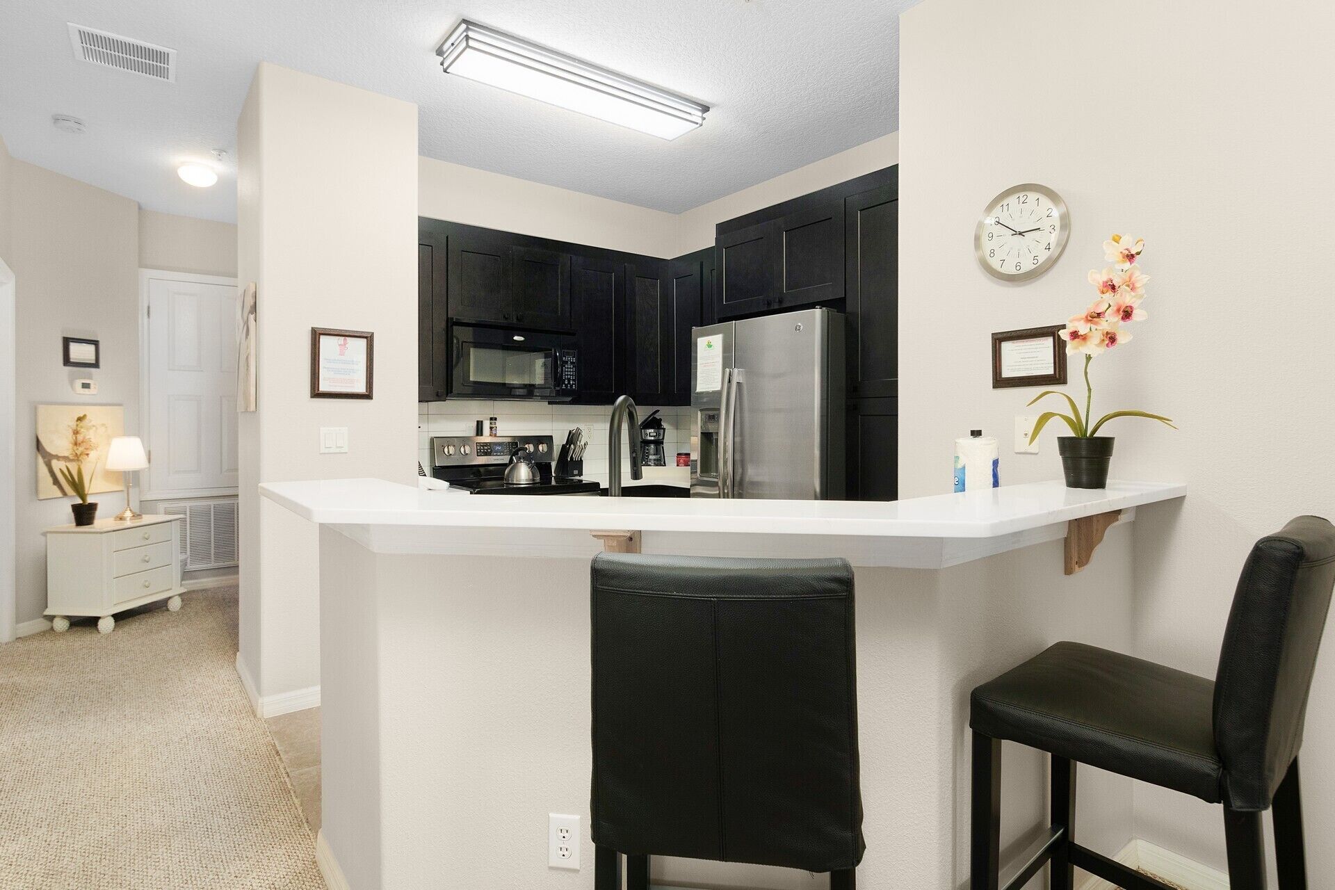 A compact kitchen with dark cabinets, a stainless steel refrigerator, a microwave, and a sink. There are two black bar stools and a small potted orchid on the counter.