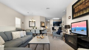 A modern living room with a gray sofa, a coffee table, and a TV. There is a dining area with chairs and a table, and a kitchen in the background. - Lakeside-Beautiful 3 bed Oakwater condo over the lake, just 1.5 miles from WDW! (Kissimmee)
