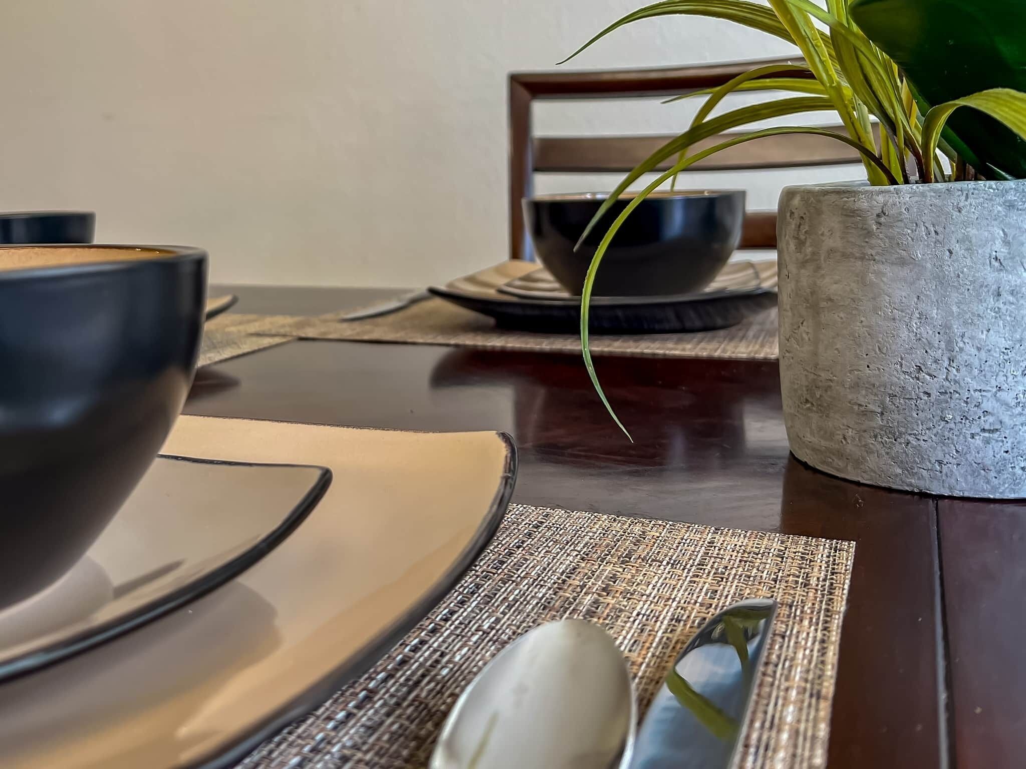 Une table à manger avec un plateau en bois à la teinte foncée, un set de table en fibres tressées, une cuillère blanche et une plante en pot aux longues feuilles vertes.