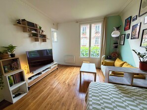 Ein Wohnzimmer mit Flachbildfernseher, Holzboden, einer Couch, einem Couchtisch und einem Fenster mit Blick auf Gebäude.