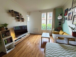 A living room with a TV, wooden flooring, a sofa, a table, and a window with a view of buildings.