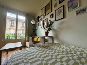 A bedroom with a checkered bedspread, a desk with a chair, a potted plant, and a window with a view of buildings.