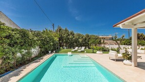 Una piscina con agua cristalina azul, rodeada por un jardín con diversas plantas y un área de estar exterior blanca con sillones reclinables.
