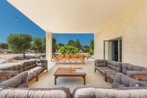 Eine Terrasse im Freien mit einem Holztisch, gepolsterten Sitzgelegenheiten und Blick auf Bäume sowie ein Gebäude im Hintergrund.