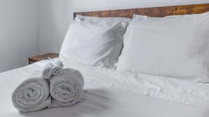 Een net opgemaakt bed met witte lakens en twee kussens, een houten hoofdbord en een klein nachtkastje ernaast.