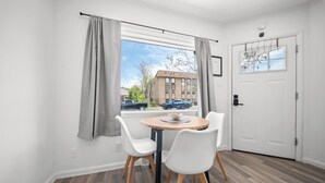 Een kamer met een ronde tafel, twee witte stoelen, een raam met gordijnen, een witte deur en uitzicht op een gebouw.