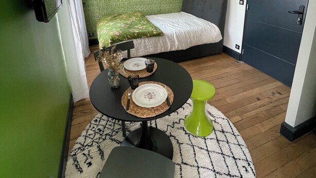 Une petite pièce comprenant un lit, une table à manger avec des assiettes et des couverts, une chaise, un tabouret vert, un miroir rond et un luminaire suspendu.