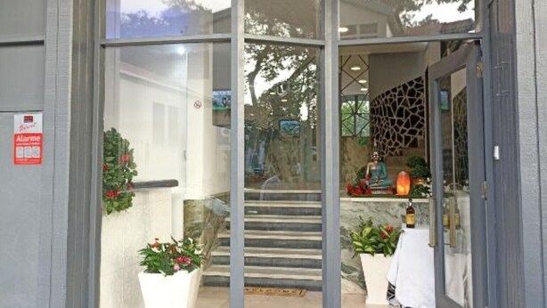 A hotel entrance with a modern design, featuring a glass door, a staircase leading inside, and a visible reception area with a decorative wall and plants.