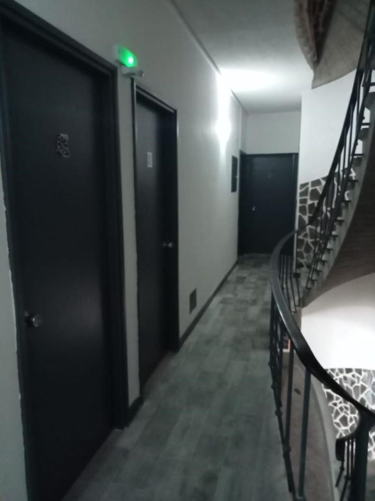 A hallway with a carpeted floor, a staircase with a metal railing, and a door with a green light above it.