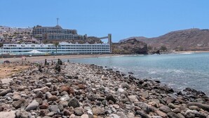 Un hôtel situé sur la côte, avec une plage rocheuse, un pont et une chaîne de montagnes en arrière-plan.