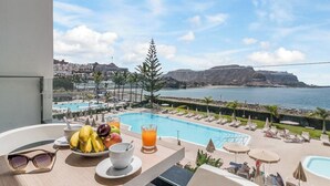 Un balcon avec vue sur une piscine, des chaises longues et une table à manger où sont disposés des fruits et des boissons.