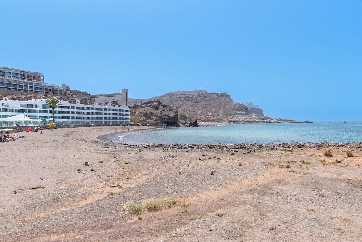 A beach with a rocky shoreline, a large building with multiple balconies, and a prominent rocky hill in the background.