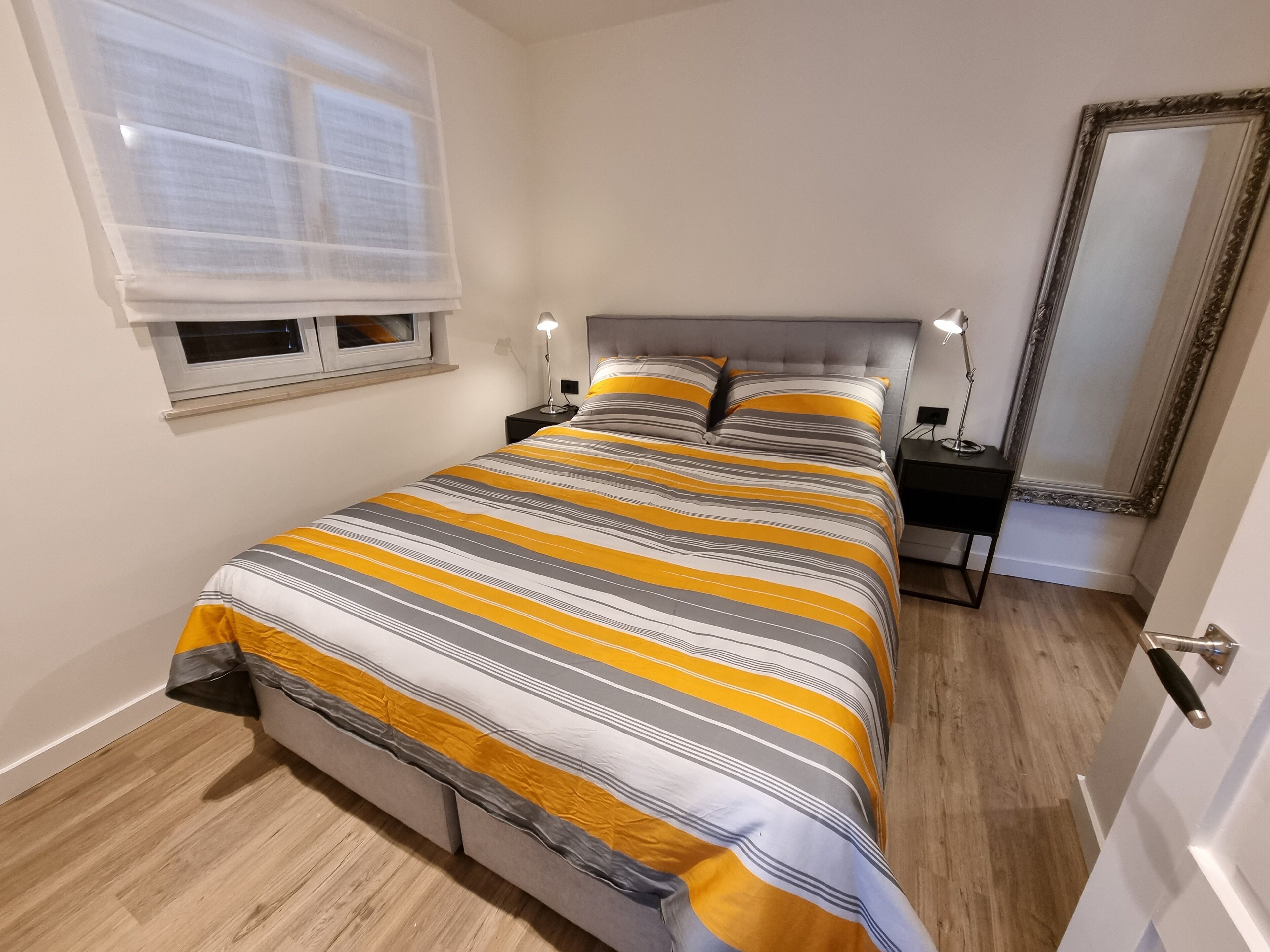 A neatly arranged bedroom with a bed, bedside lamps, a window with blinds, a mirror, and a door.