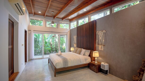Een slaapkamer met een groot bed, houten plafond en uitzicht op groen door de ramen.