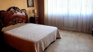 Une chambre dotée d’un grand lit, d’une tête de lit en bois, d’une petite table de chevet avec une lampe et d’une fenêtre ornée de rideaux légers.