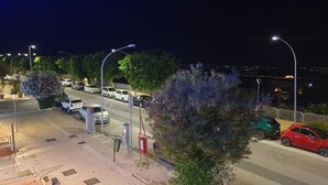 Une vue nocturne d’une rue, avec des voitures garées, des lampadaires et un pont en arrière-plan.