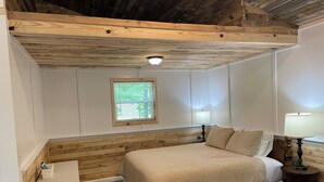 Une chambre avec un plafond en bois, un lit avec une literie beige, une table de chevet avec une lampe, une fenêtre avec des stores et une petite armoire blanche.