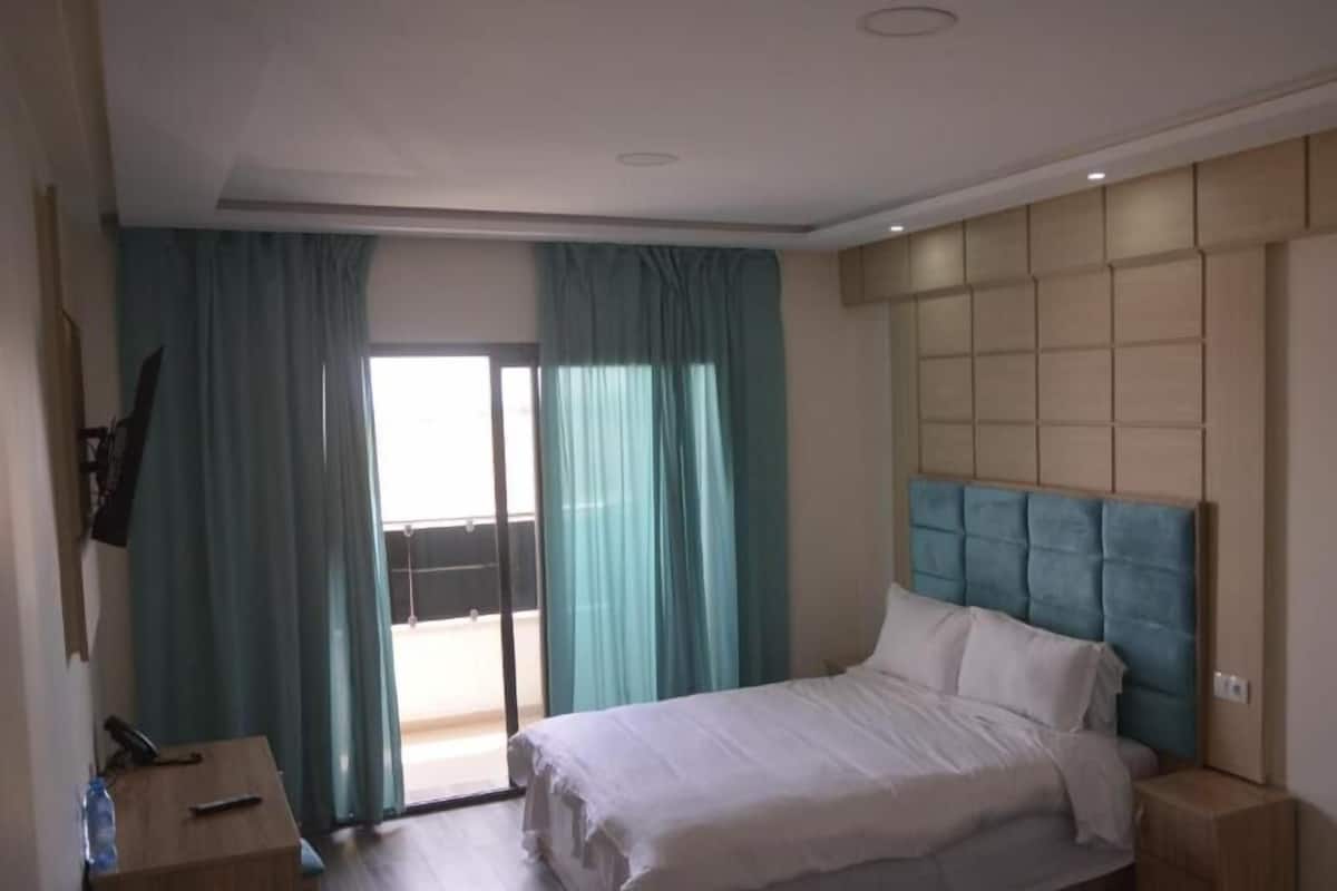 A hotel room with a bed, a small table, a TV mounted on the wall, and a view through the window.
