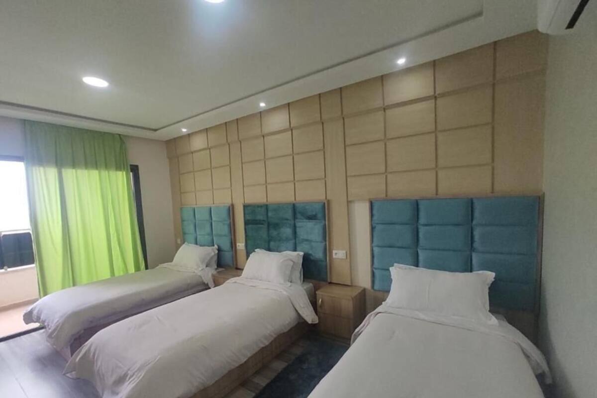 A hotel room with three beds, a large window with green curtains, and a modern headboard design.