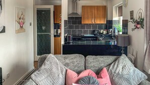 A living room with a grey sofa, pink cushions, and a floral pillow. A kitchen with dark tiles and wooden cabinets is visible in the background. - Sunny Rhyl Apartment - UK44631 (Rhyl)