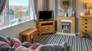 A cozy living room with a fireplace, a television on a wooden stand, and a window with curtains. - Sunny Rhyl Apartment - UK44631 (Rhyl)
