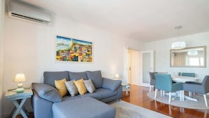 Ein modernes Wohnzimmer mit grauem Sofa, passendem Hocker, Essbereich mit Tisch und Stühlen sowie einem großen Bild an der Wand.
