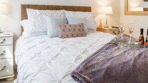Een netjes opgemaakt bed met witte beddengoed en een bruin hoofdbord, een nachtkastje met een lamp, en een dienblad met twee glaasjes en een fles.