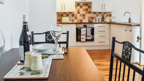 Een eettafel met een fles wijn, glazen en een bord voor twee personen. Achtergrond toont een keuken met witte kasten en een geruit spatwand.