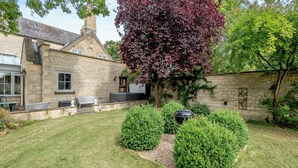 A stone house with a chimney, a barbecue area, and a well-maintained garden with hedges and a tree with red leaves.