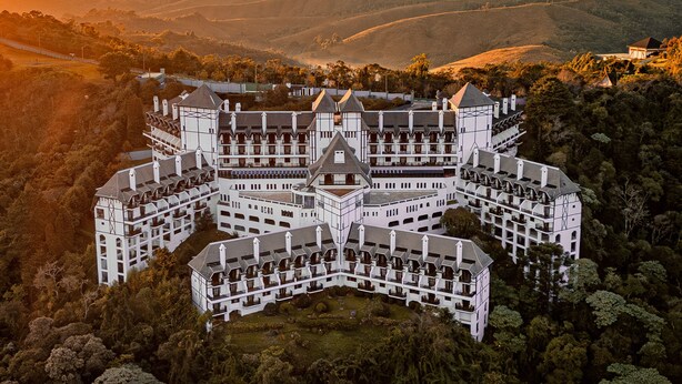 Un grand bâtiment symétrique, doté de nombreuses ailes et d’une tour centrale, entouré d’une forêt dense et de collines.