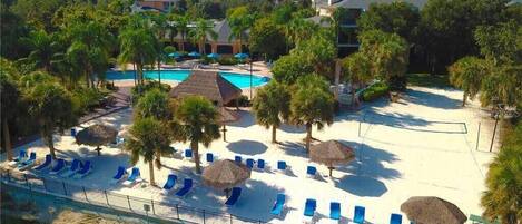 An aerial view of a resort with a swimming pool, beach area with sun loungers, and palm trees.
