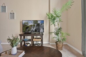 Una sala de estar con televisor sobre un mueble de madera, una mesa de centro, una planta en maceta y una estantería.