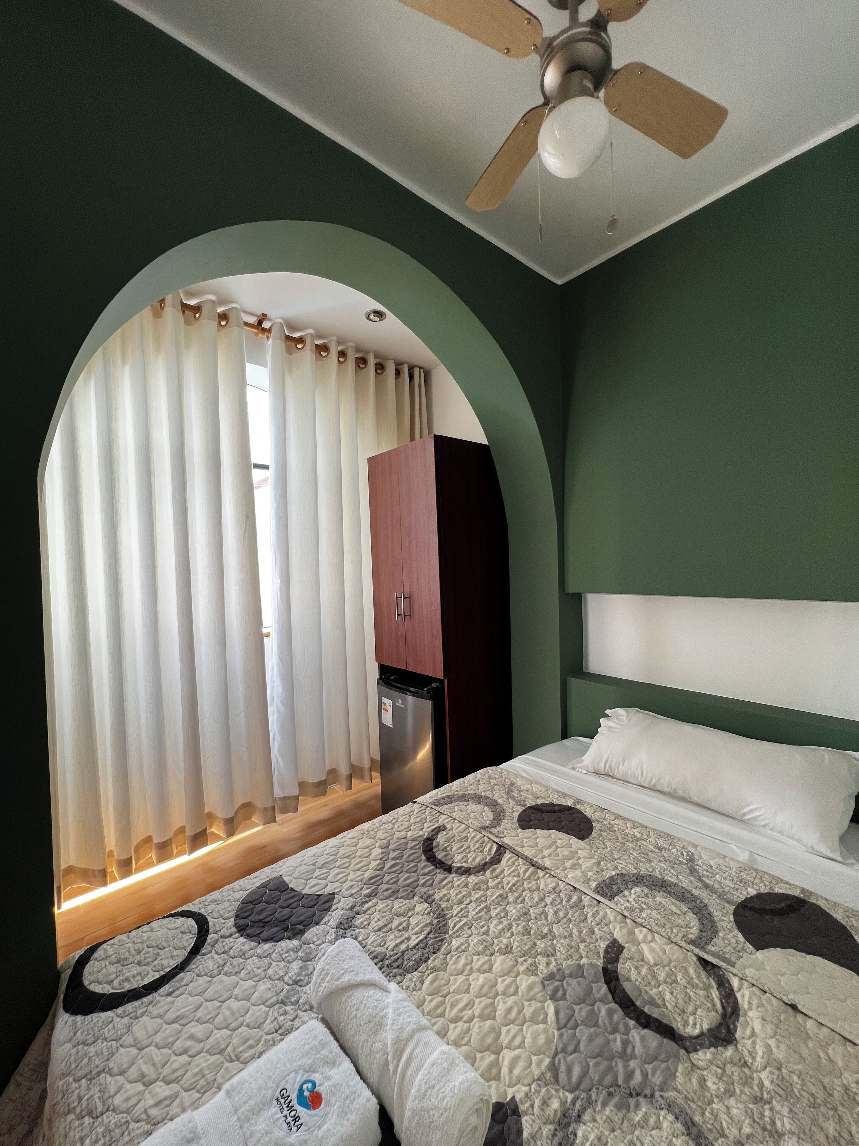 Ein Schlafzimmer mit Bett, Deckenventilator, einem Fenster mit Vorhängen und einer Tür zu einem Badezimmer.