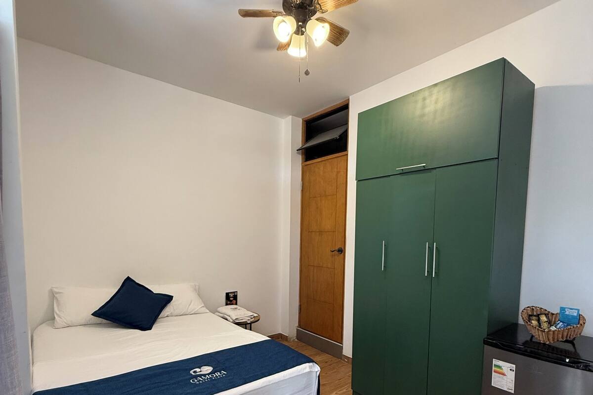 Una camera d'albergo con un letto, una ventola a soffitto, un armadio e un tavolino con una lampada.