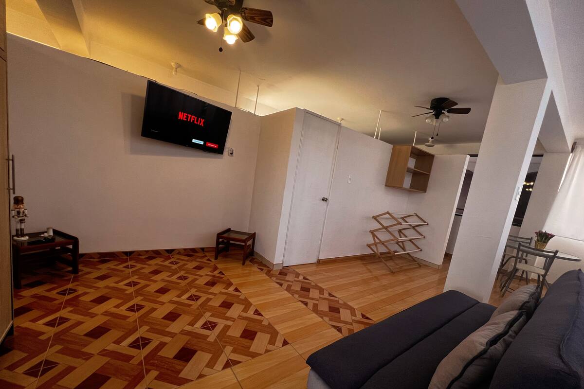 Un soggiorno moderno con pavimento in legno decorato, una TV a muro che mostra Netflix, una ventola a soffitto e un divano.