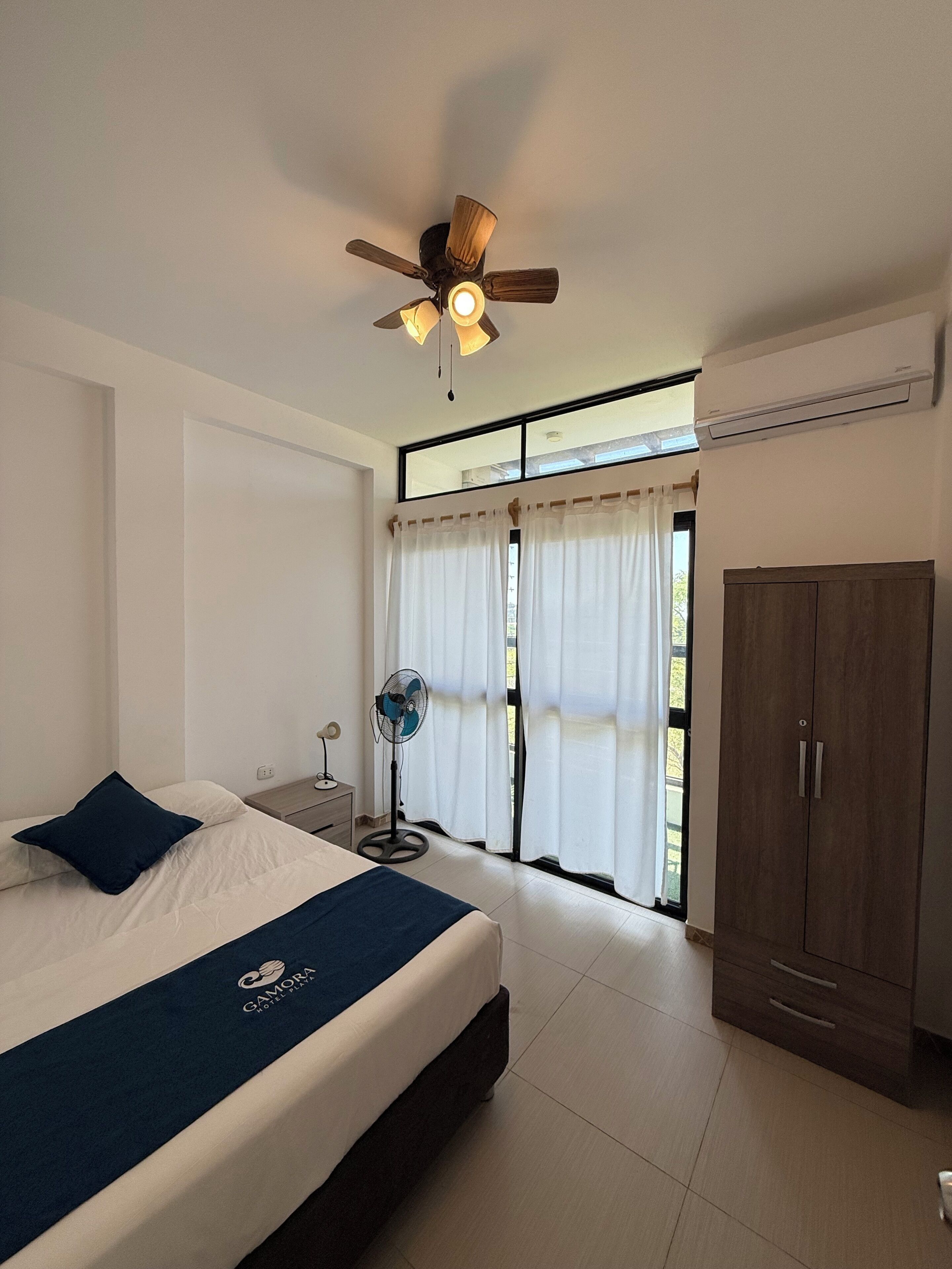 A bedroom with a bed, a ceiling fan, a standing fan, a wardrobe, and a window with curtains.