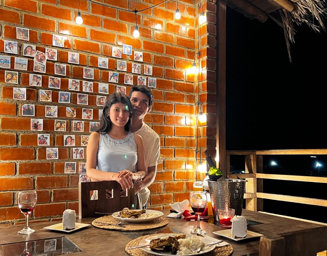 A couple dining at a table with a brick wall adorned with photos, a bottle of champagne, and a bucket of ice.