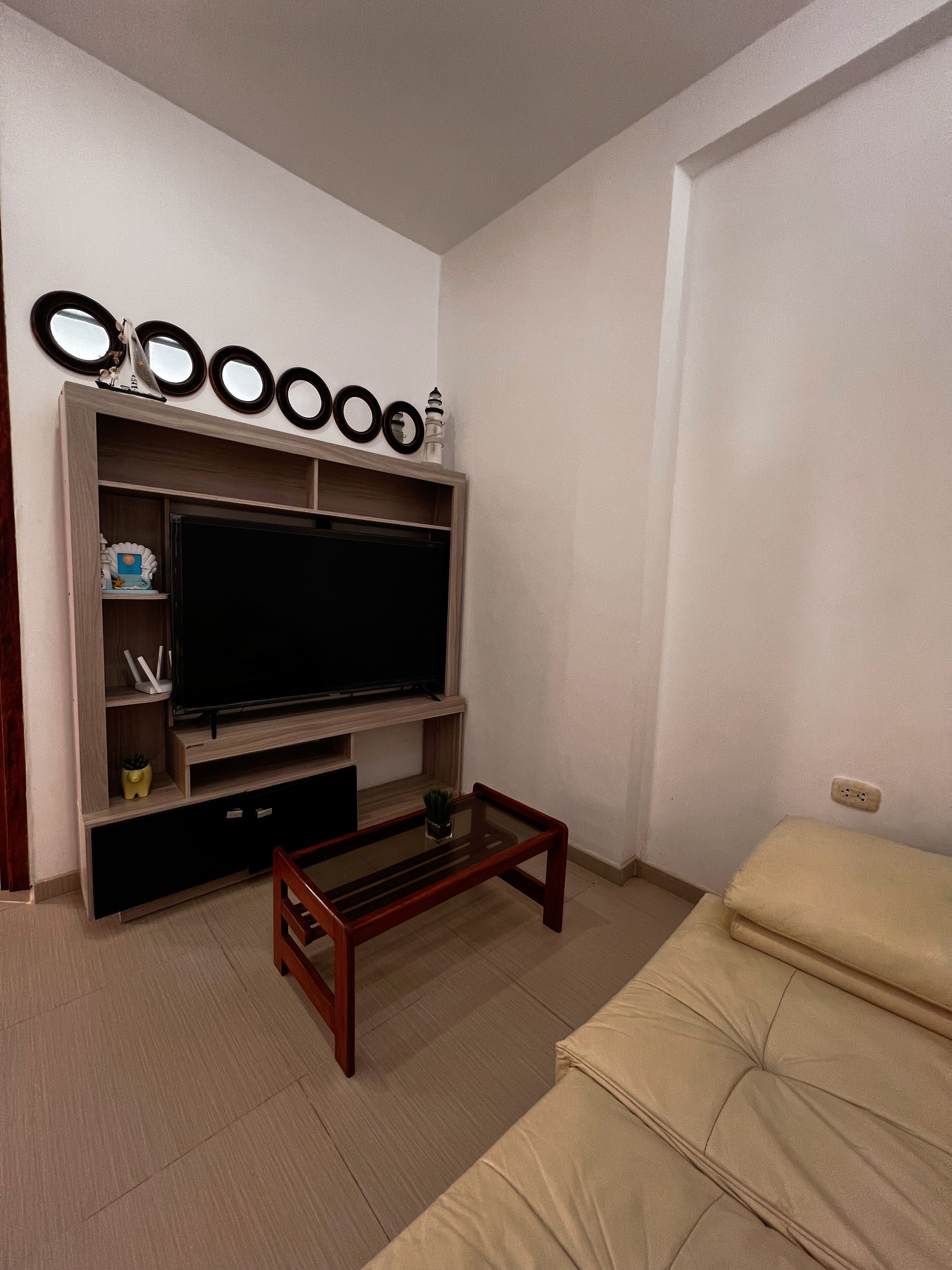 Ein Wohnzimmer mit Flachbildfernseher auf einem Holzbodenständer, einem Glastisch und einem beigefarbenen Sofa.