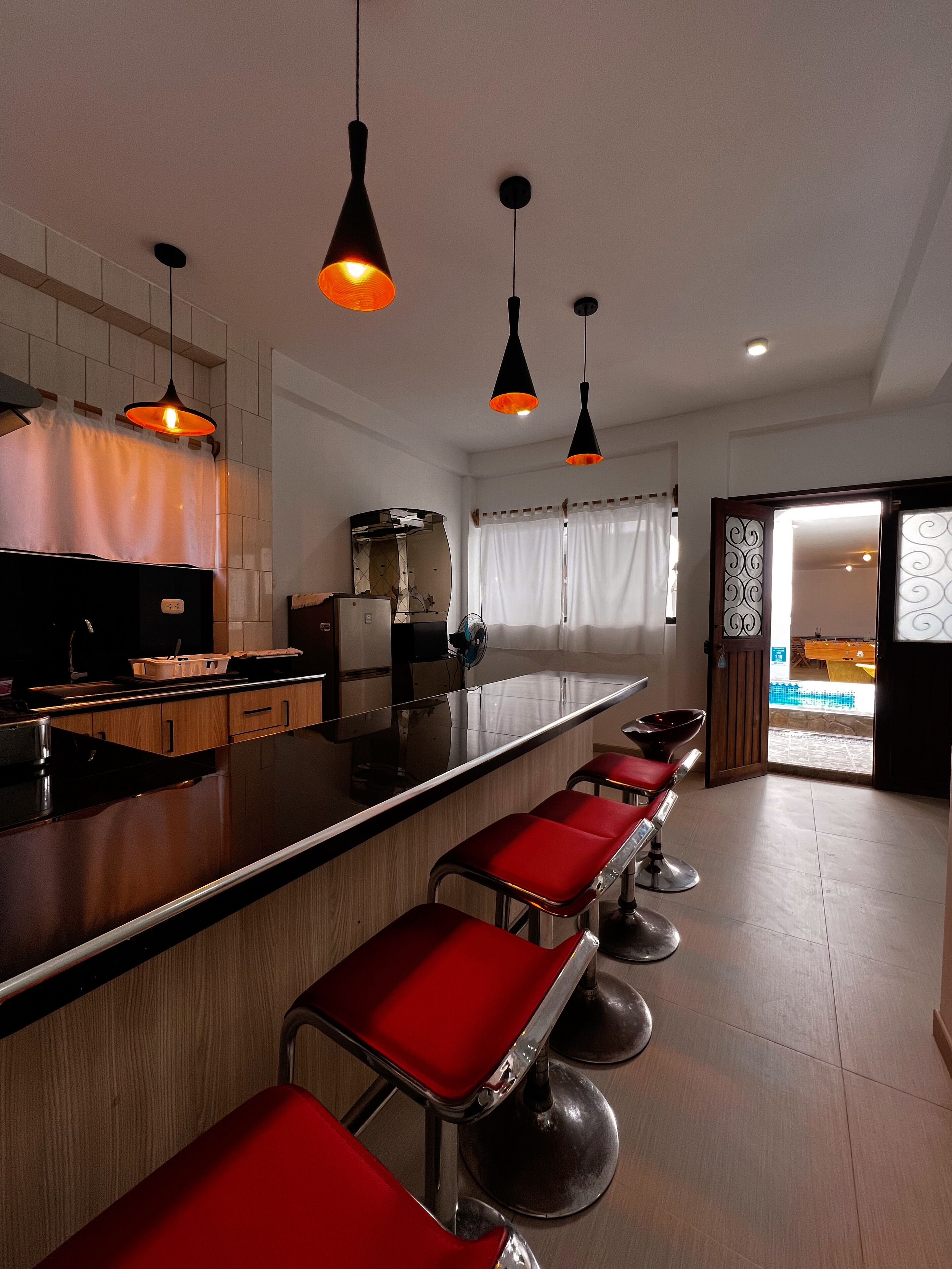 Cocina moderna con una barra alargada, taburetes rojos, iluminación colgante y vistas a un área de piscina exterior.