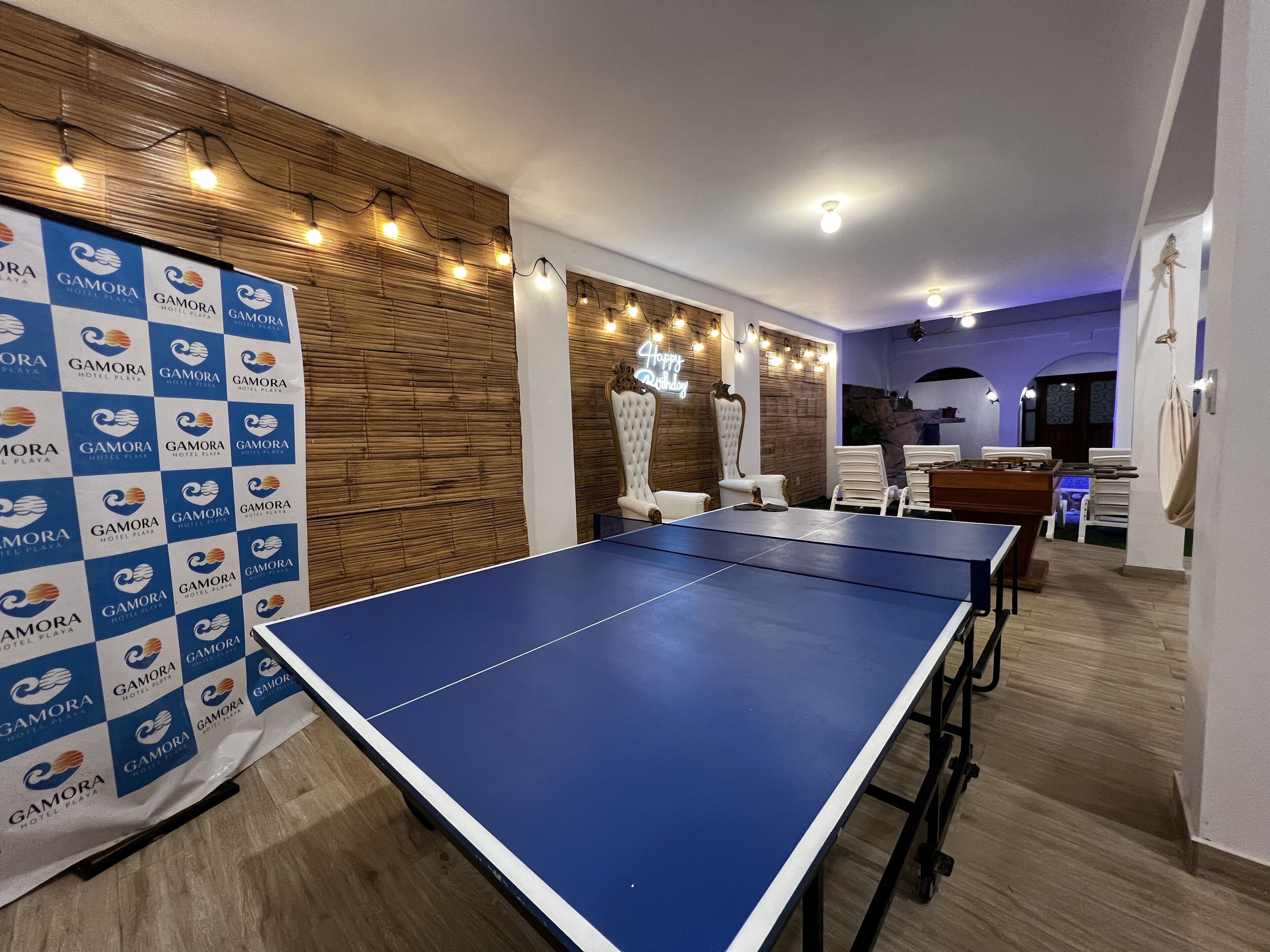 A room with a ping pong table, white chairs, a wooden wall, and a banner with the logo of Gamora Hotel Playa.