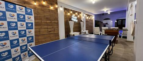 A room with a ping pong table, white chairs, a wooden wall, and a banner with the logo of Gamora Hotel Playa.